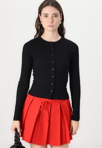 Čierny rebrový kardigán s dlhým rukávom, okrúhlym výstrihom a prednými gombíkmi, skombinovaný s plisovanou červenou sukňou a čiernou taškou.