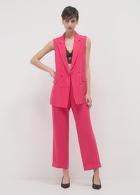 Knallpinker Zweiteiler mit einem ärmellosen Blazer mit doppelreihigen Knöpfen und weit geschnittenen Hosen. Texturierte Spitzen-Bralette sichtbar darunter.