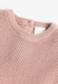 Suéter de bebé de punto en suave color rosa con un patrón texturizado y un cuello redondo acanalado sobre un fondo blanco.
