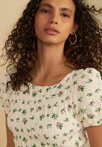 Haut floral blanc avec des manches bouffantes, comprenant des feuilles vertes et des fleurs roses en tissu texturé. Encolure ronde, élastiqué à la taille.