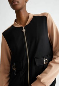 Giubbotto bomber nero con maniche nude, caratterizzato da una chiusura con zip, due tasche davanti, dettagli in hardware dorato e tessuto texture.