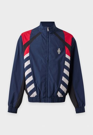 Un mannequin masculin se tient devant un fond uni, vêtu d’une casquette marron, d’une veste de survêtement bleu marine avec des empiècements rouges et blancs, d’une chemise beige, d’un jean clair et de bottes noires.