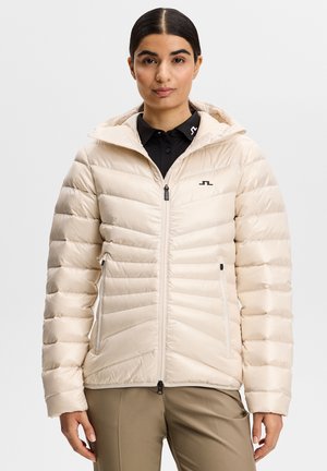 Kvinde iført en cremefarvet, quiltet dunjakke med lynlås foran og sidelommer, kombineret med khakifarvede bukser og en sort skjorte med krave.