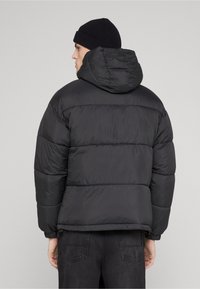 Svart dunjakke med hette, som har horisontale vatterte seksjoner, elastiske mansjetter og en ledig passform. Brukt over mørke jeans.