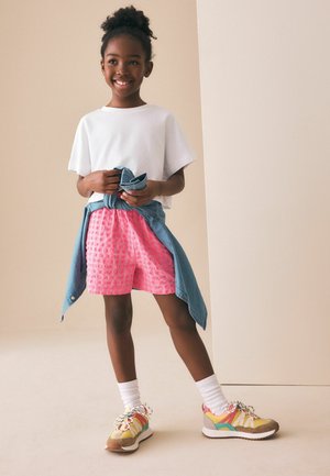 Naeratav tüdruk seisab, kandes valget t-särki, roosad tekstuuriga lühikesed püksid, kollased tossud ja teksariidest jopet, mis on tal vöö ümber seotud, beeži tausta ees.