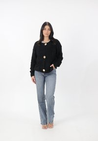 Maglione nero e morbido con tre bottoni dorati, tasche frontali e una vestibilità rilassata, abbinato a jeans azzurri e tacchi trasparenti.