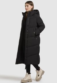 Schwarzer langer Daunenmantel mit Kapuze, ausgestattet mit gestepptem Futter und seitlichen Taschen. Trägt dazu schwarze Leggings und beige klobige Stiefeletten.