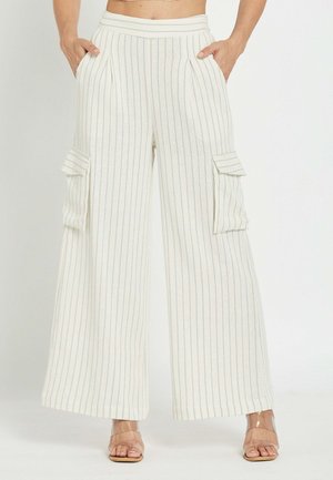 Belo bele hlače z visokim pasom in širokimi hlačnicami, z ozkimi navpičnimi črtami in velikimi žepi s poklopcem na straneh, nošene z odprtimi petami.