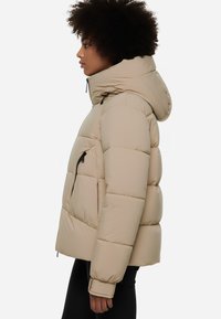 Giacca puffy beige con cappuccio, caratterizzata da un design trapuntato, tasca laterale con zip e polsini elasticizzati. Texture liscia e materiale leggero.