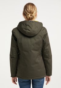 Kvinna med blont hår i en knut, iklädd en mörkgrön jacka med huva och blå jeans, sedd bakifrån mot en neutral bakgrund.