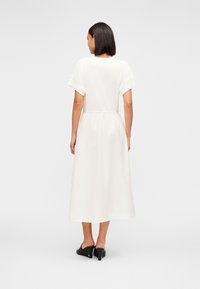 Λευκό μακρύ φόρεμα με κοντά μανίκια, ζώνη στη μέση και φαρδιά φούστα. Λείο ύφασμα με διακριτική γυαλάδα; Συνδυασμένο με μαύρες γόβες.