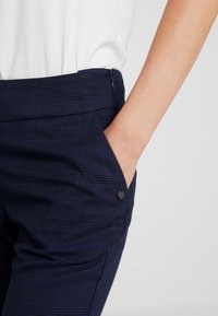 Námořnické kárované kalhoty s hladkým povrchem, které mají boční kapsu a dekorativní černý knoflík na pase.