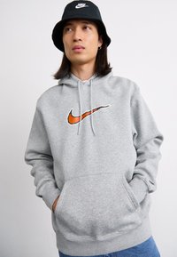Grå hættetrøje med stort Nike swoosh-logo i orange og sort. Kængurulomme, snor i hætten, blanding af bomuldsstof. Båret sammen med blå jeans.
