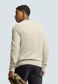 Licht beige textuurgebreide trui met geribde manchetten en zoom, gecombineerd met donkere broeken, model houdt een bruine geblokte shirt vast.