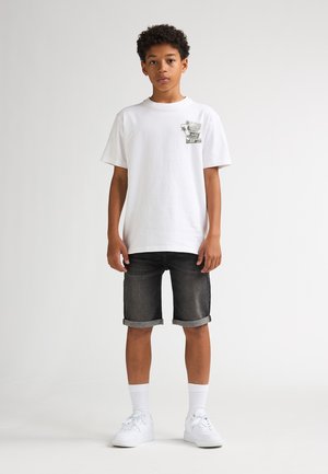 T-shirt en coton blanc avec un motif vert sur la poitrine, accompagné de shorts en denim foncé, de baskets blanches et de chaussettes blanches montantes.