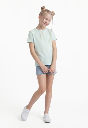 Jente med flettehår og hårboller, iført en lyseblå t-skjorte, denimshorts og hvite joggesko, smilende og stående med korslagte ben.