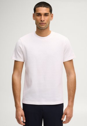 Mann mit kurzen Haaren, der ein hellrosa strukturiertes Kurzarmhemd und dunkle Hose trägt, steht vor einem einfarbigen Hintergrund.