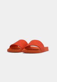 Paire de sandales slides orange à bout ouvert avec larges brides en tissu et semelles rembourrées sur un fond clair uni.