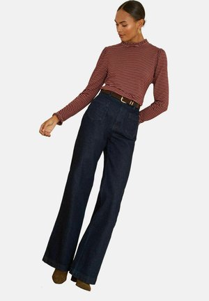 Raidallinen punavalkoinen pitkähihainen paita, jossa on röyhelöhiharesorit, yhdistettynä tummansinisiin korkeavyötäröisiin flared-farkkuihin ja ruskeaan vyöhön, käyttäjänä ruskeat kengät.