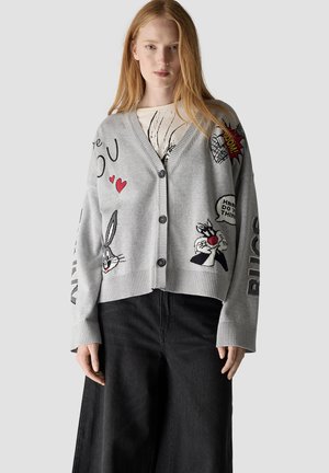 Woman wearing a gray buttoned cardigan with cartoon characters and text, paired with a white shirt and black wide-leg pants.