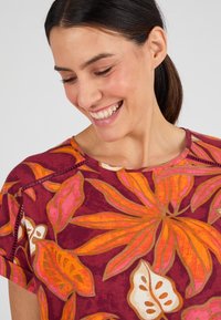 Top a maniche corte realizzato in un vivace tessuto bordeaux con grandi motivi floreali arancioni e rosa, e cuciture decorative vicino al collo.