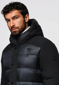 Zwarte pufferjack met een hoge kraag, horizontaal gestepeld patroon en matte zwarte accenten. Voorzien van drukknoopsluiting en een rits.
