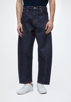 Persona che indossa jeans a gamba larga di colore blu scuro e sneakers bianche, in piedi davanti a uno sfondo semplice.
