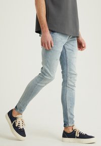 Ljust blå skinny jeans med slitdetaljer, kombinerat med marinblå sneakers med beiga snören och vita sulor. Bärs med en grå skjorta.