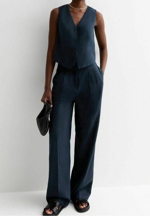 Woman wearing a sleeveless dark blue buttoned vest and matching wide-leg pants, holding a black woven handbag, with black sandals.