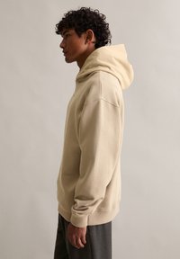 Beżowa bluza z dużym, miękkim kapturem. Wykonana z teksturowanej tkaniny, o luźnym kroju i ściągaczach przy rękawach. Prosty design, bez logo.