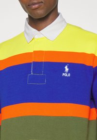 Polo shirt a righe colorate con strisce orizzontali in giallo, arancione, blu e verde, con colletto bianco e dettaglio del logo.