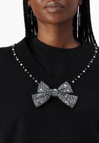 Czarny sweter z dekoltem w serek ozdobiony rzędami srebrnych cyrkonii wzdłuż linii dekoltu i dużą zdobioną biżuterią kokardą na środku.