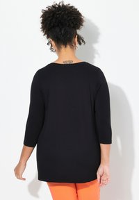Maglietta nera a maniche lunghe con scollatura rotonda e fit rilassato, realizzata in tessuto morbido e con un sottile dettaglio di tatuaggio sulla schiena. Indossata con pantaloni arancioni.