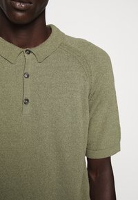 Olivově zelené polo tričko vyrobené z texturované pleteniny, s límečkem a nápletem na tři knoflíky. Krátké rukávy s žebrovanými manžetami.