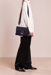 Kobieta w białej bluzce i czarnych spodniacach o szerokich nogawkach, nosząca granatową torebkę z złotym zapięciem oraz czarne czółenka z ćwiekami.