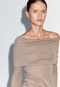 Frau mit slicked-back Haar, die ein beiges, schulterfreies Strickoberteil trägt und gegen einen schlichten, hellen Hintergrund schaut.