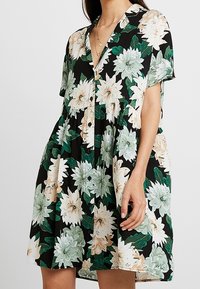 Blommig klänning i svart med gröna och krämfärgade blommor, korta ärmar, en knäppning framtill och en lös, flödande form. Slät tygtextur.