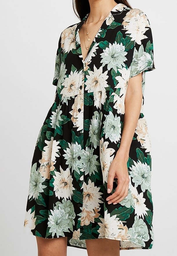 Blommig klänning i svart med gröna och krämfärgade blommor, korta ärmar, en knäppning framtill och en lös, flödande form. Slät tygtextur.