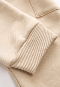 Tejido de sudadera de algodón beige con detalle de puños acanalados, que presenta una textura suave y un acabado blando. Sin patrones visibles ni piezas metálicas.