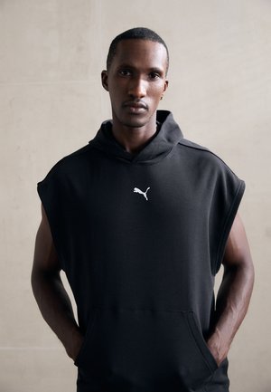Mann mit Händen in den Taschen trägt eine ärmellose schwarze Puma-Hoodie und steht an einer schlichten hellen Wand.