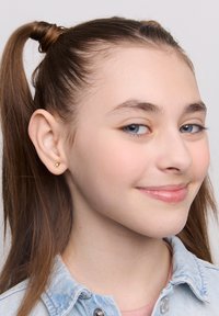 Złoty kolczyk w kształcie serca, mały i gładki, noszony na jasnoskórej uchu. Jasnobrązowe włosy ułożone w dwa kucyki. Jasnoniebieska koszula dżinsowa.