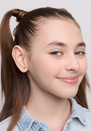 Złoty kolczyk w kształcie serca, mały i gładki, noszony na jasnoskórej uchu. Jasnobrązowe włosy ułożone w dwa kucyki. Jasnoniebieska koszula dżinsowa.