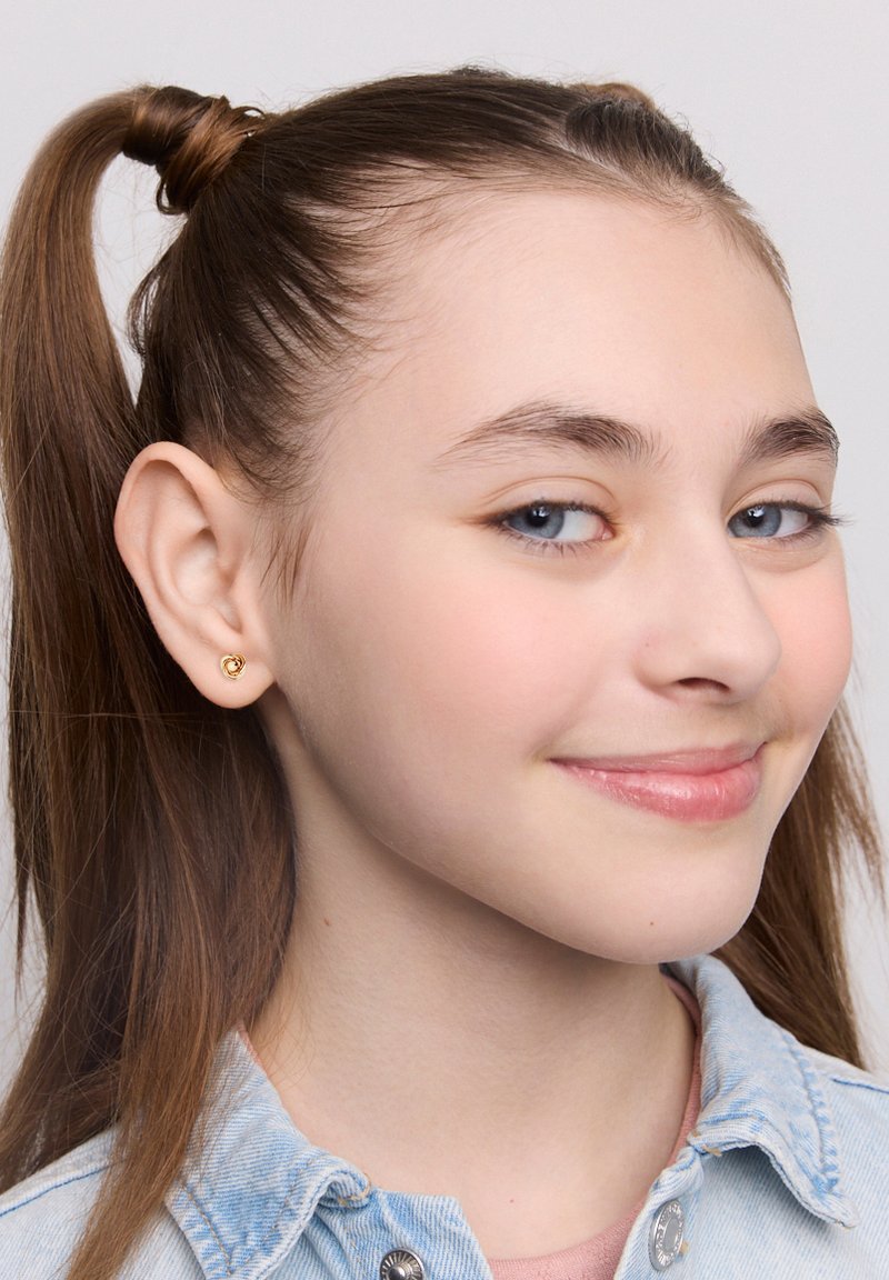 Złoty kolczyk w kształcie serca, mały i gładki, noszony na jasnoskórej uchu. Jasnobrązowe włosy ułożone w dwa kucyki. Jasnoniebieska koszula dżinsowa.