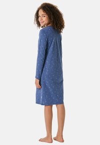 Meisje met krullend haar, gekleed in een blauwe nachtjapon met lange mouwen en sterrenpatronen, staand op blote voeten en over haar schouder kijkend.