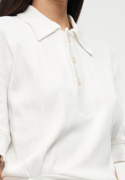 Polo blanc à côtes avec col classique et patte à trois boutons. Comprend des manches courtes et un tissu doux et texturé.