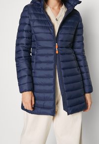 Meriharmaa puhvitakki, jossa on vetoketjullinen etuosa, huppu ja vaakasuora tikkikuvio. Varustettu oranssilla vetoketjunvedolla ja pehmeällä, sileällä pinnalla.