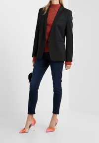 Blazer negro, cuello alto de rayas rojas, jeans de mezclilla oscura y tacones de punta multicolor con tacón de aguja. Sin accesorios notables.
