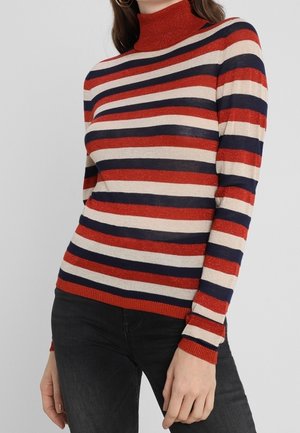 Vrouw draagt een aangesloten coltrui met horizontale strepen in rood, marineblauw en beige, gecombineerd met een zwarte broek.