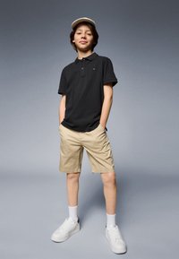Polo nero, pantaloni khaki, scarpe da ginnastica bianche, con un cappellino chiaro. Tessuto liscio e vestibilità casual. Postura eretta.