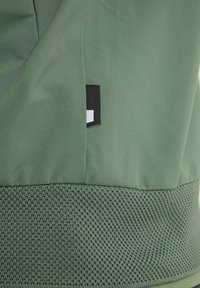Gros plan d’une veste en tissu vert avec une ceinture en tricot texturé et une petite étiquette noire et blanche sur une couture.